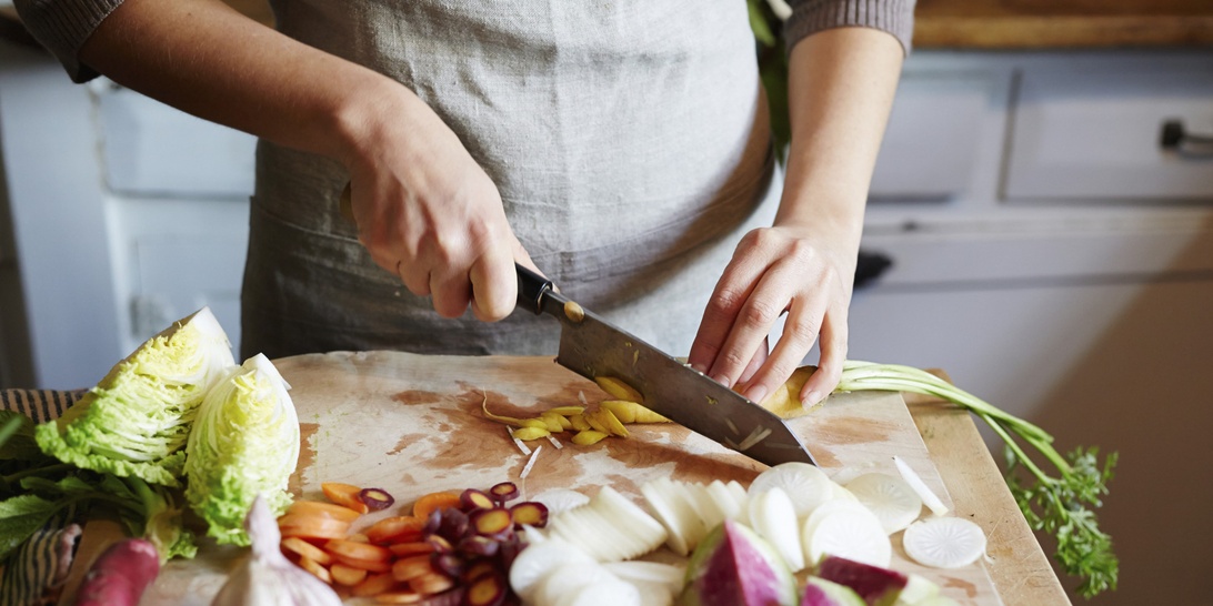 Närbild på händer som hackar färska grönsaker på en träskärbräda, omgiven av förportionerade ingredienser typiska för matkassar.